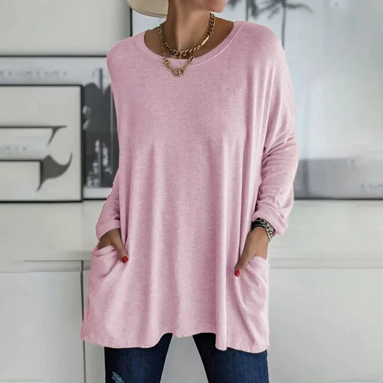 Femme portant un pull rose ample avec poches, collier doré, et jeans. Style décontracté chic, mode féminine tendance, intérieur moderne.