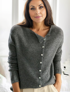 Femme portant un cardigan gris en laine avec boutons, posant devant une fenêtre lumineuse. Mode automne-hiver, style décontracté et élégant.