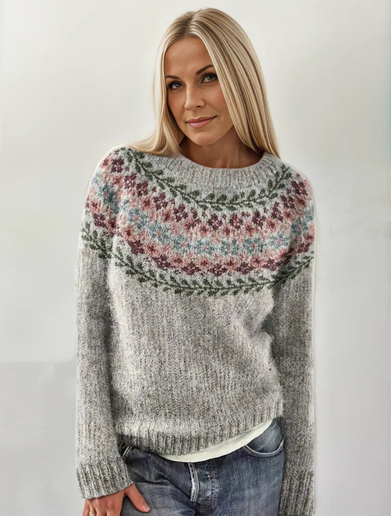 Femme portant un pull en laine gris avec motif nordique coloré, cheveux blonds lisses, posant devant un fond neutre. Mode hiver, style scandinave.