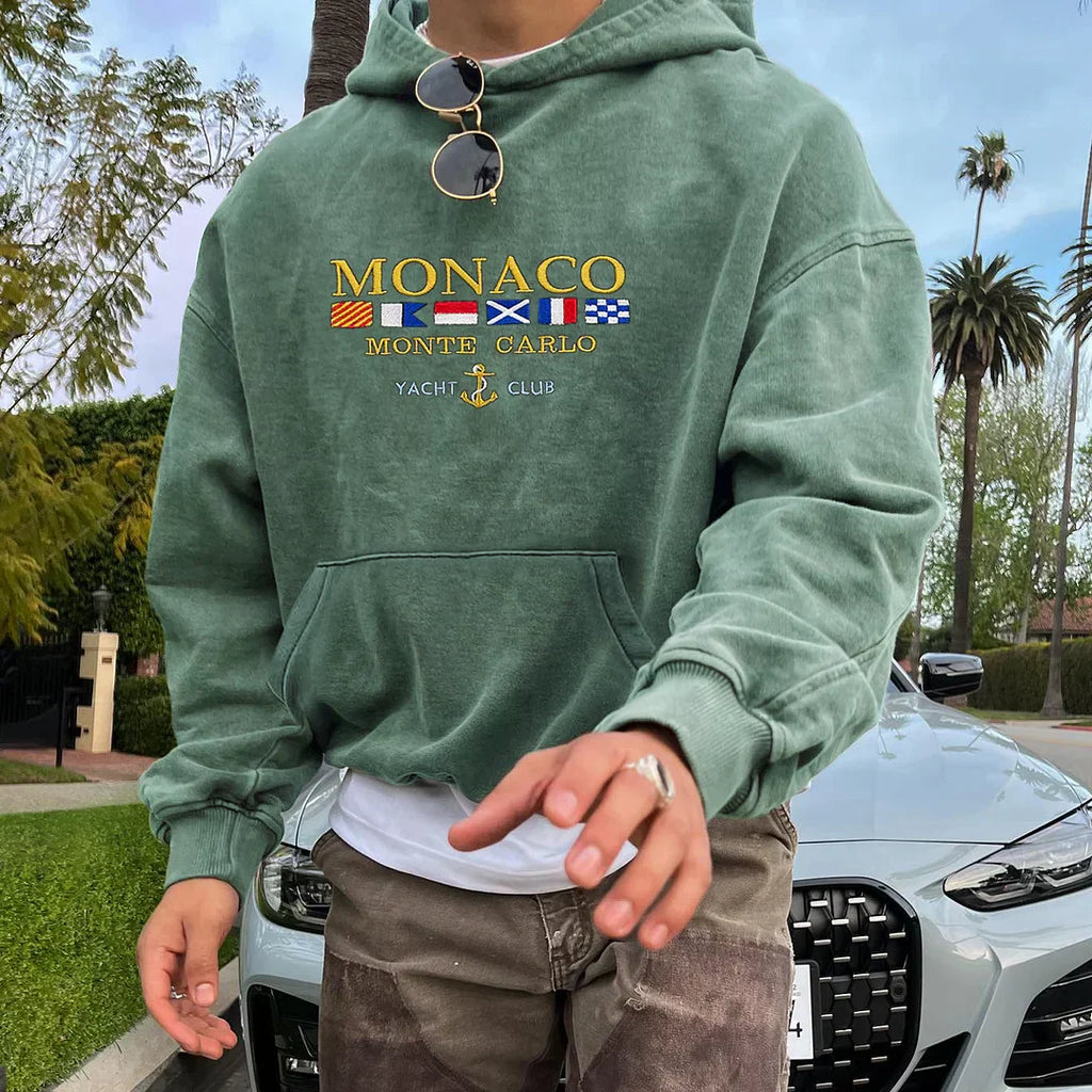 Homme portant un sweat à capuche vert "Monaco Monte Carlo Yacht Club" devant une voiture de luxe, entouré de palmiers. Mode décontractée, style urbain.