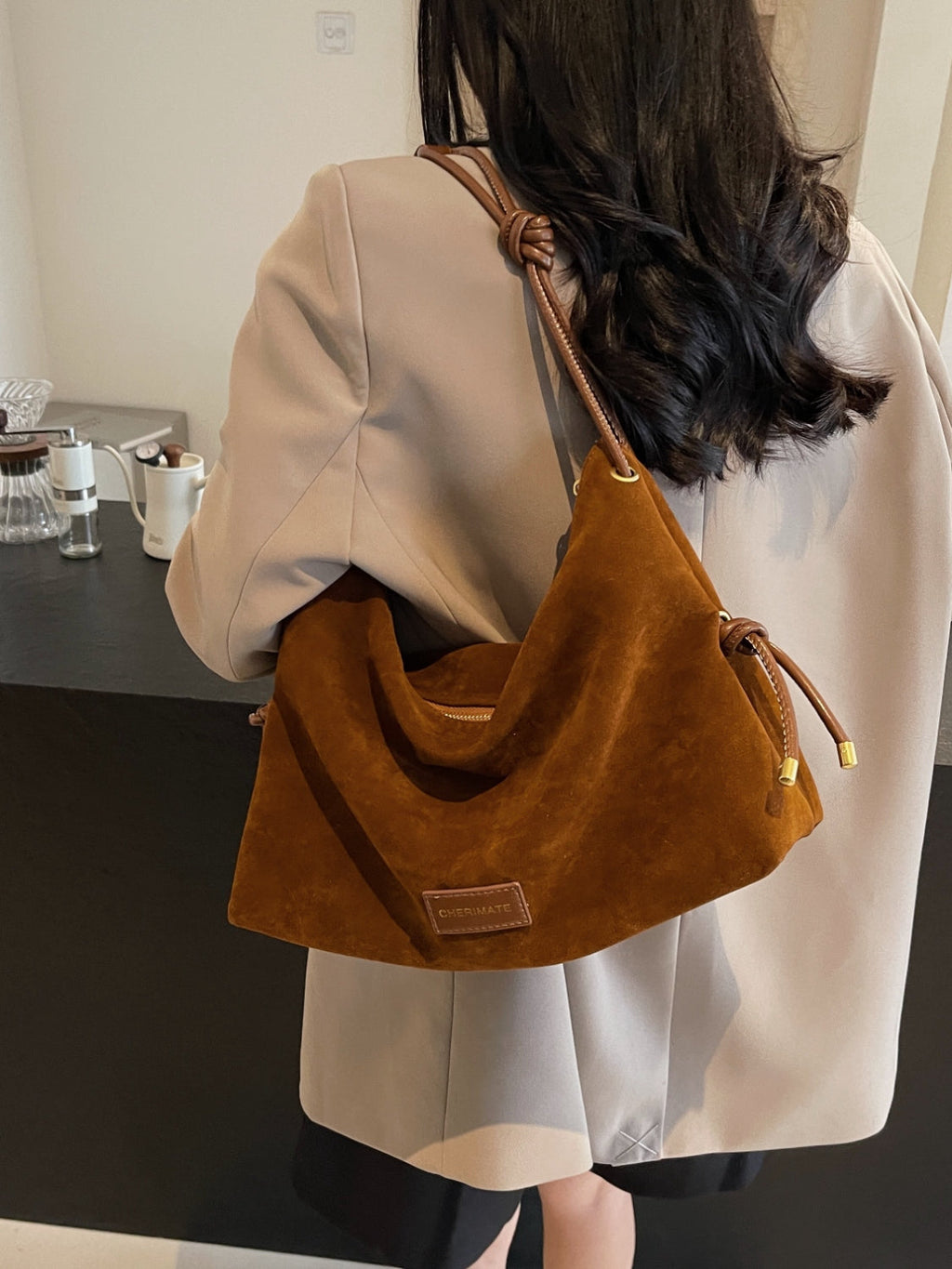 Femme portant un sac à main en daim marron, élégant et moderne, avec une étiquette en cuir, sur fond de comptoir noir. Accessoire mode tendance.
