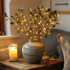 Vase en céramique avec branches lumineuses LED, posé sur une table en bois à côté d'une bougie allumée, créant une ambiance chaleureuse et cosy.