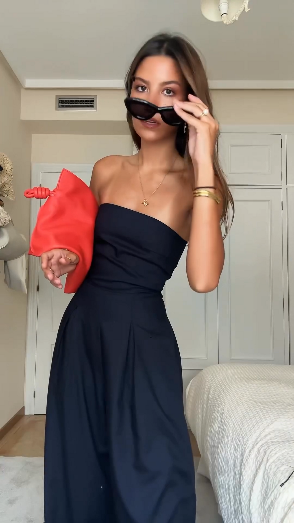 Jeune femme en robe noire élégante, tenant une pochette rouge, ajustant ses lunettes de soleil. Style chic, mode féminine, accessoires tendance.