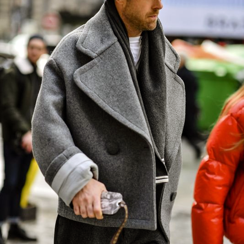Homme portant un manteau gris oversize, style streetwear, avec écharpe noire, tenant un sac transparent. Mode urbaine, tendance hiver.