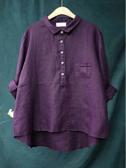 Chemise en lin violet pour femme, manches longues retroussées, col boutonné, poche poitrine, suspendue sur un cintre en bois, fond vert.