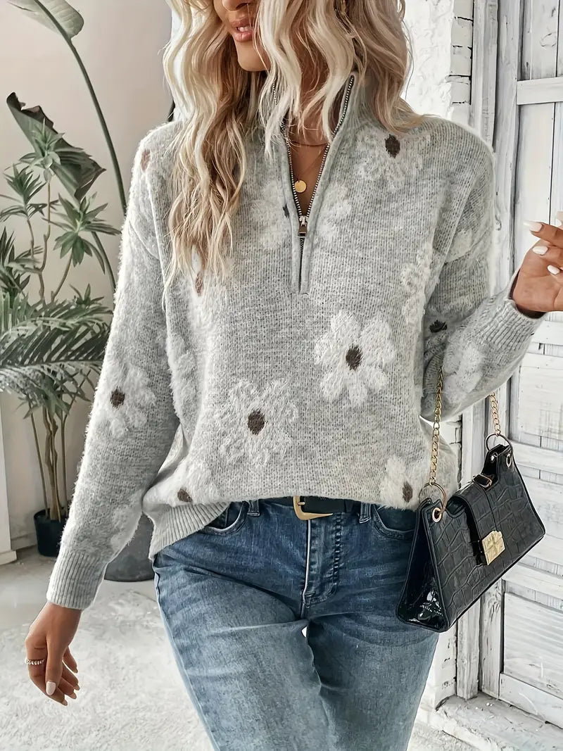 Femme portant un pull gris à fleurs blanches, jeans décontracté, sac à main noir, cheveux blonds ondulés, intérieur moderne avec plantes.