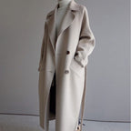 Manteau long beige élégant pour femme, style automne-hiver, avec col large et boutons, porté sur un pull blanc à col roulé.