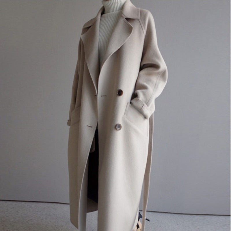 Manteau long beige élégant pour femme, style automne-hiver, avec col large et boutons, porté sur un pull blanc à col roulé.