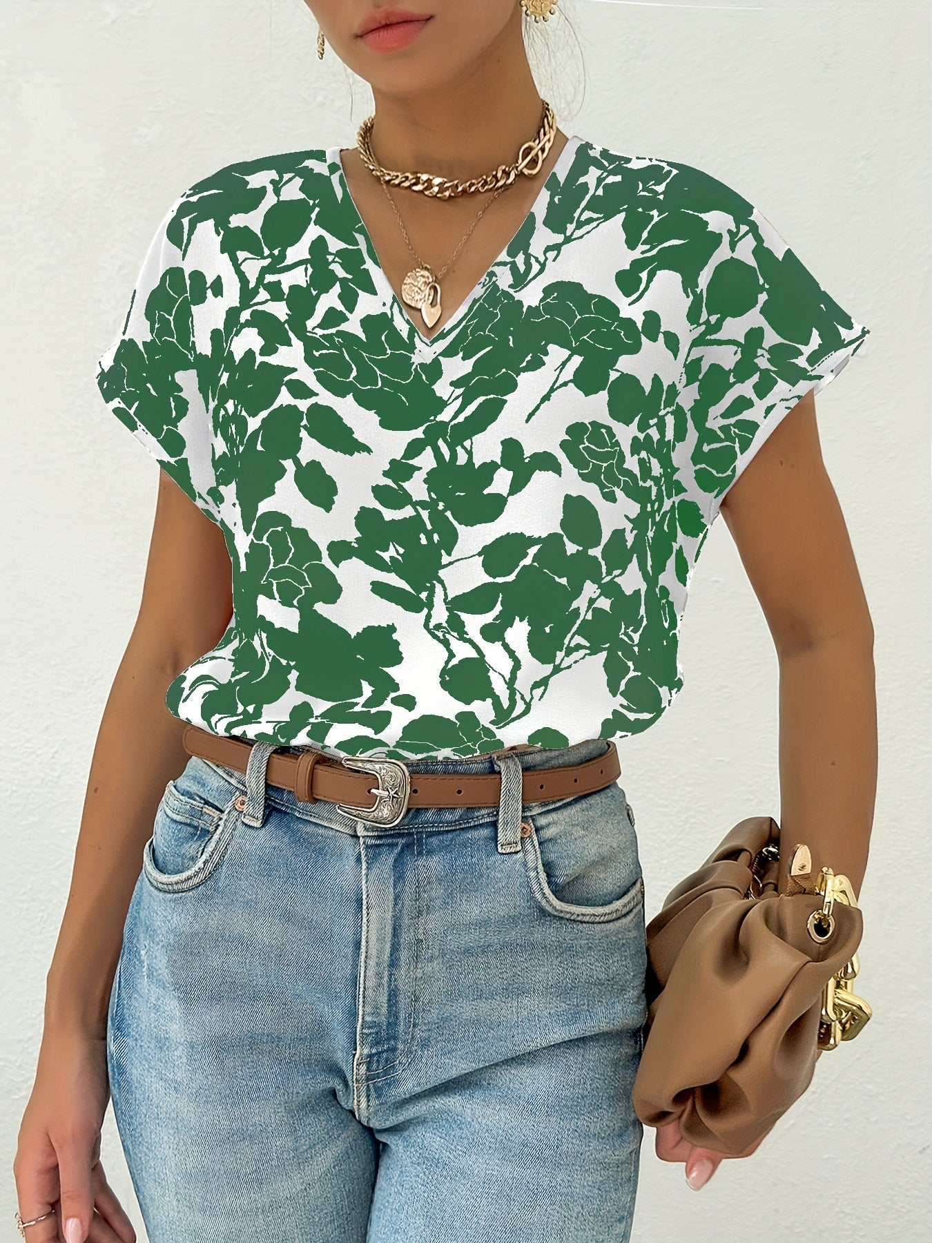 Femme portant un t-shirt à motifs floraux verts et blancs, jean bleu, ceinture marron, sac à main beige, collier doré. Mode décontractée élégante.