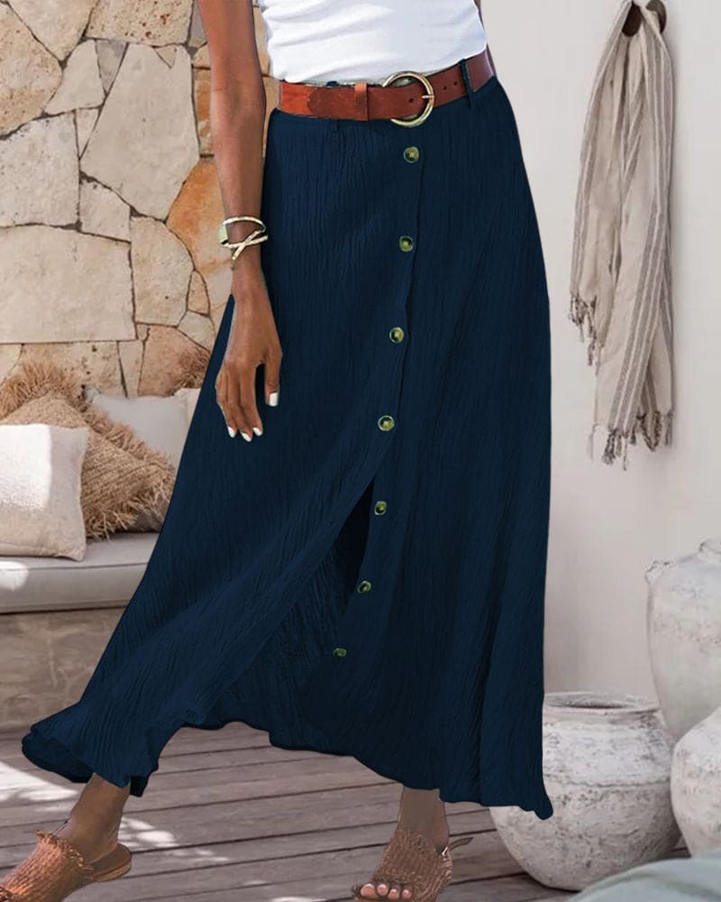 Robe longue bleue à boutons, ceinture marron, femme debout sur terrasse en bois, décor rustique avec coussins et poteries. Mode estivale élégante.