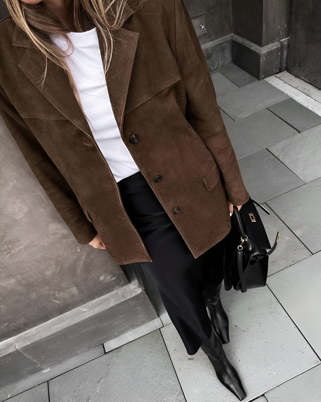 Femme en manteau marron en daim, jupe noire et bottes, tenant un sac à main noir. Mode automne chic, style urbain élégant.