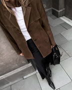 Femme en manteau marron en daim, jupe noire et bottes, tenant un sac à main noir. Mode automne chic, style urbain élégant.