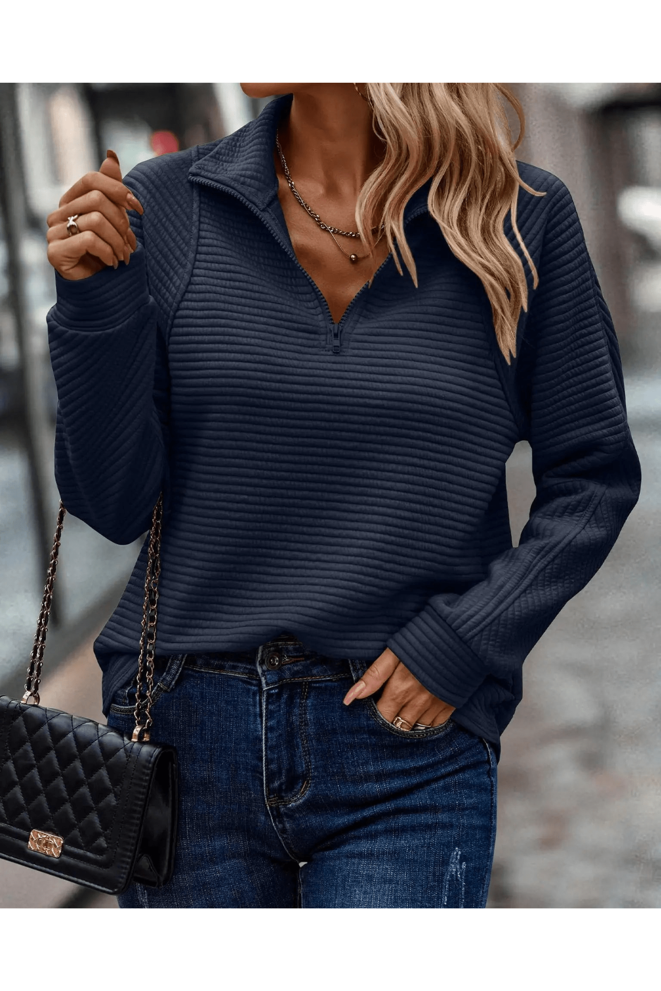 Femme portant un pull bleu marine à col zippé, avec un sac à main noir matelassé et un jean, marchant en ville. Mode automne-hiver élégante.