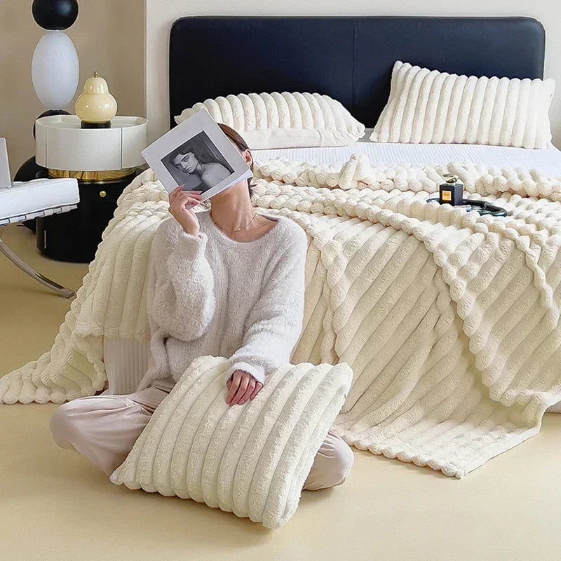 Femme en pull blanc assise sur un lit avec couverture et oreillers en peluche crème, tenant une photo. Décor moderne, ambiance cosy et élégante.
