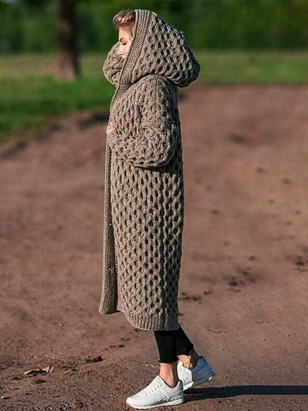 Femme portant un long manteau en tricot beige avec capuche, debout sur un chemin de terre. Mode automne, style confortable, vêtements chauds.