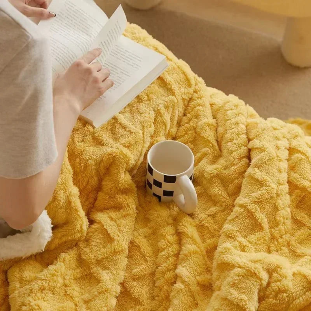 Personne lisant un livre sur une couverture jaune moelleuse avec une tasse à carreaux noirs et blancs, ambiance cosy et détente à la maison.