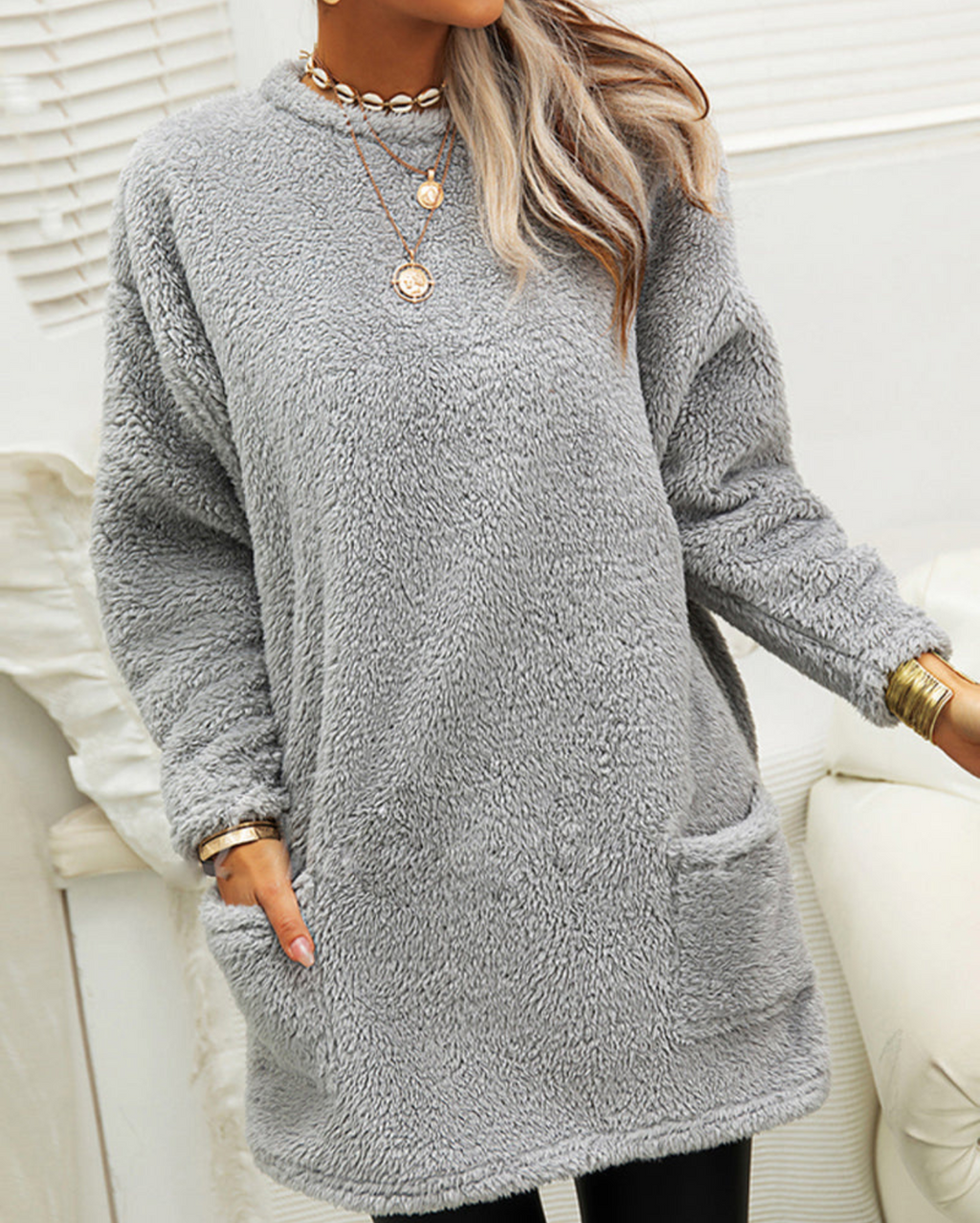 Femme portant un pull en peluche gris avec col rond, manches longues et poches avant. Style décontracté, idéal pour l'hiver. Accessoires dorés visibles.