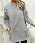 Femme portant un pull en peluche gris avec col rond, manches longues et poches avant. Style décontracté, idéal pour l'hiver. Accessoires dorés visibles.