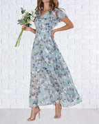 Robe longue fleurie bleue, manches courtes, femme tenant un bouquet de fleurs blanches, fond mur en briques blanches, élégance estivale.