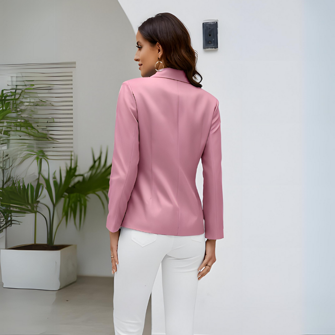 Femme de dos portant une veste rose élégante et un pantalon blanc, posant dans un intérieur moderne avec des plantes vertes. Mode féminine chic.