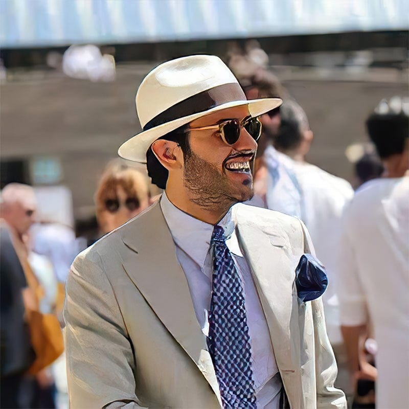 Homme élégant en costume beige, cravate bleue, chapeau panama blanc et lunettes de soleil, souriant lors d'un événement en plein air.