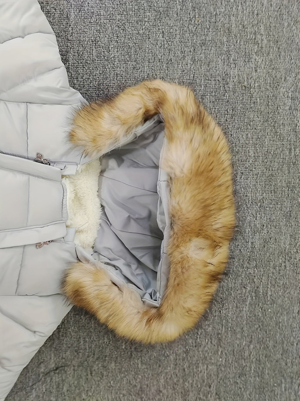 Doudoune grise avec capuche en fausse fourrure marron, posée sur un tapis gris. Veste chaude hiver, mode femme, tendance.