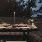 Table de dîner en plein air au crépuscule avec lampe moderne, assiettes blanches, verres à vin et décorations florales, ambiance élégante et naturelle.