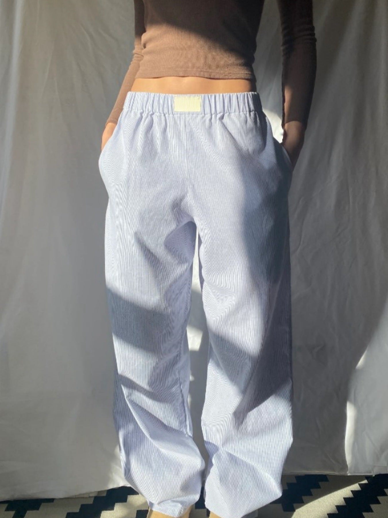 Personne portant un pull marron et un pantalon ample blanc à rayures, éclairée par la lumière naturelle. Mode décontractée, style minimaliste.