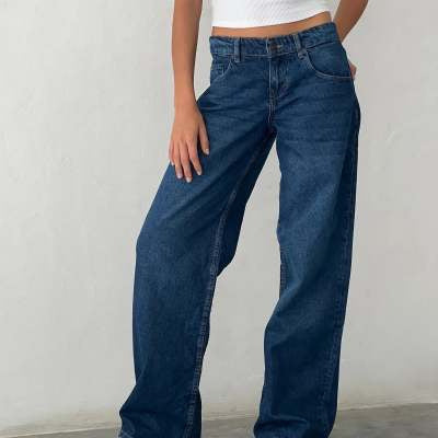 Jeune femme portant un jean large bleu et un haut blanc côtelé, posant contre un mur blanc. Mode décontractée, tendance jeans larges.