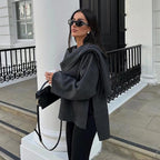 Femme élégante portant un manteau gris, lunettes de soleil noires, tenant un sac à main et une boisson chaude, devant un bâtiment blanc chic.