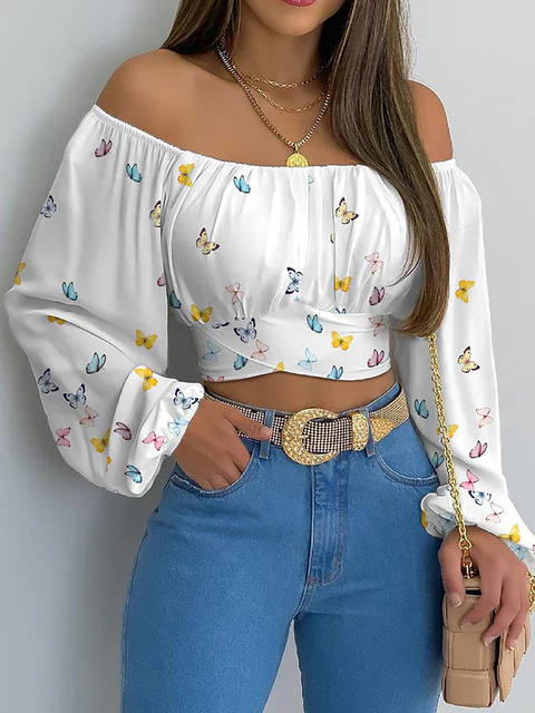 Femme portant un haut blanc à épaules dénudées avec motifs papillons colorés, jeans bleu taille haute, ceinture dorée, collier et sac à main beige.