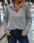 Femme portant un pull gris à col zippé et un jean bleu, posant en extérieur urbain. Mode automne, tenue décontractée, style moderne.