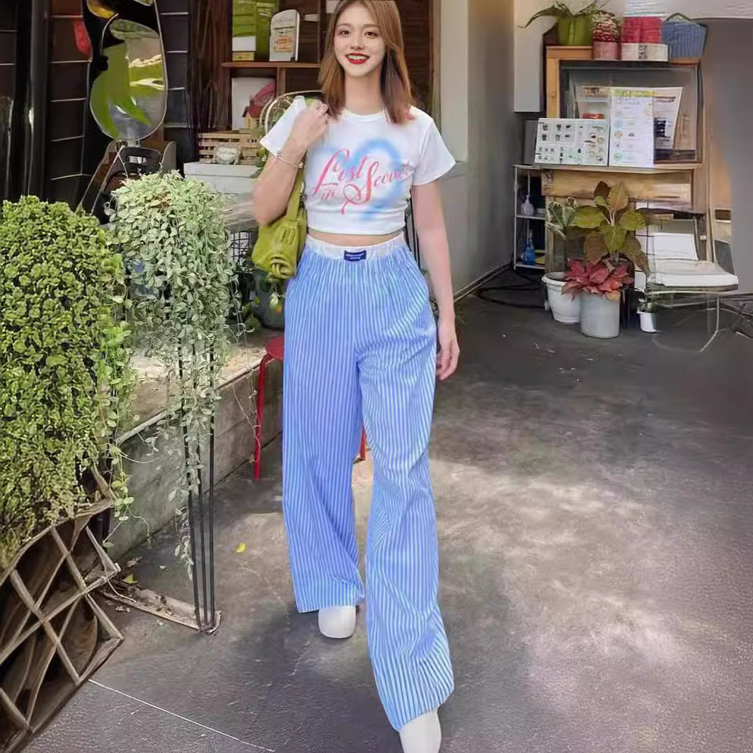 Jeune femme souriante portant un t-shirt blanc et un pantalon bleu à rayures, marchant dans une rue urbaine avec des plantes en arrière-plan.