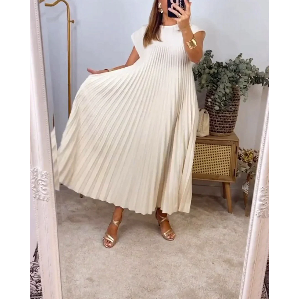 Robe plissée blanche élégante portée par une femme, posant devant un miroir. Décor minimaliste avec plantes et meuble en bois. Mode féminine chic.