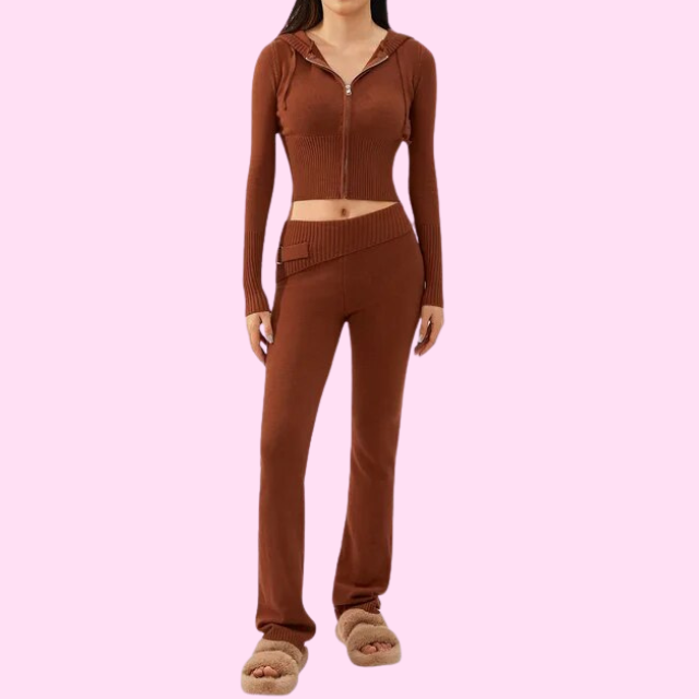 Femme portant un ensemble marron en tricot, avec un haut à capuche zippé et un pantalon taille haute, sur fond rose. Mode automne-hiver tendance.