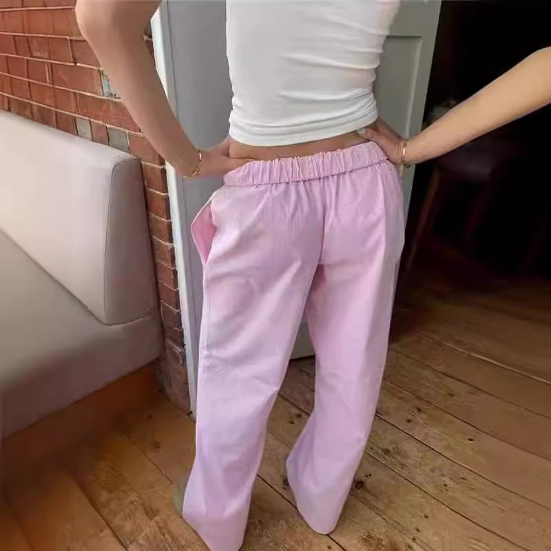 Femme portant un pantalon rose ample et un haut blanc ajusté, debout près d'un mur en briques. Mode décontractée, style estival, tenue confortable.