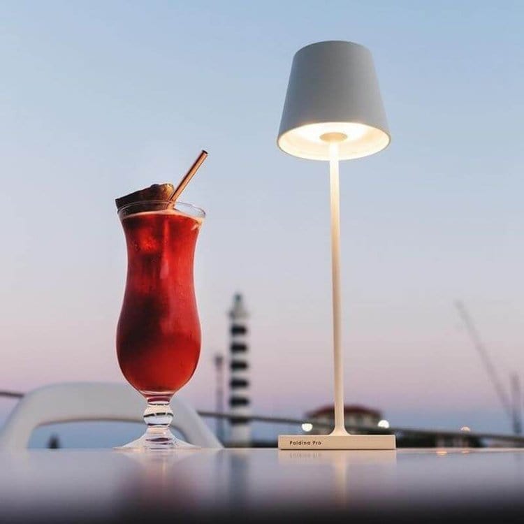 Verre de cocktail rouge avec paille et décoration, posé sur une table à côté d'une lampe moderne, ciel crépusculaire en arrière-plan.