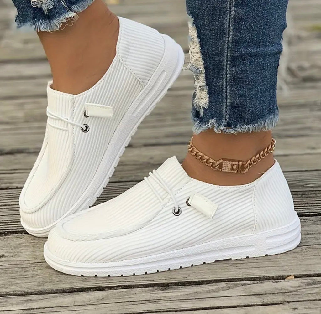 Chaussures blanches en toile côtelée, portées avec un jean déchiré et un bracelet de cheville doré, sur fond de plancher en bois.