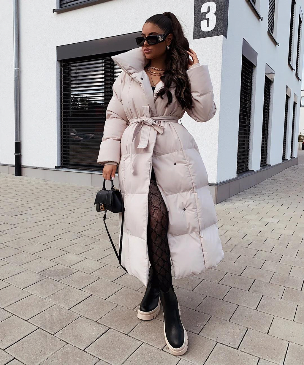 Femme portant un manteau long beige ceinturé, lunettes de soleil, bottes noires, posant devant un bâtiment moderne. Mode hiver élégante.