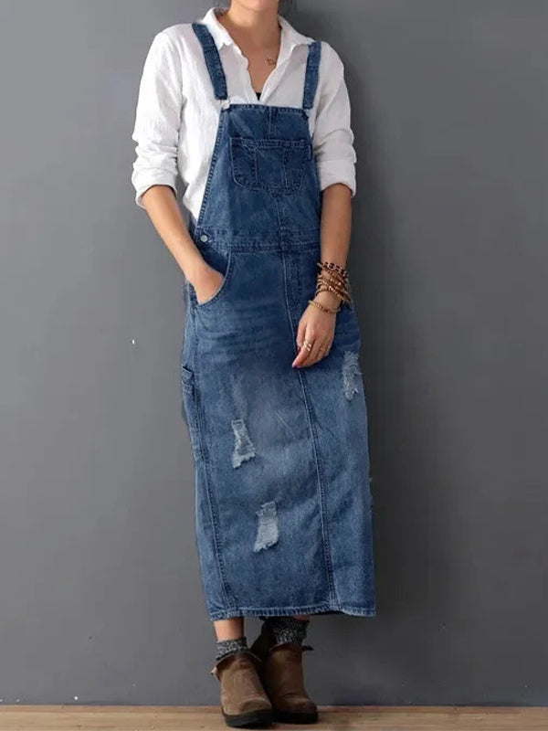 Femme portant une robe salopette en jean décontractée avec chemise blanche, posant contre un mur gris. Mode féminine, style décontracté, tendance denim.