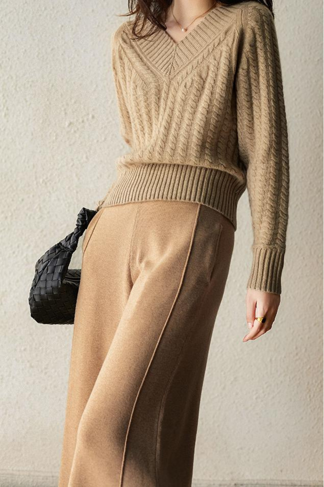 Femme en pull en maille beige et pantalon ample, tenant un sac tressé noir. Mode automne élégante, tenue décontractée chic.