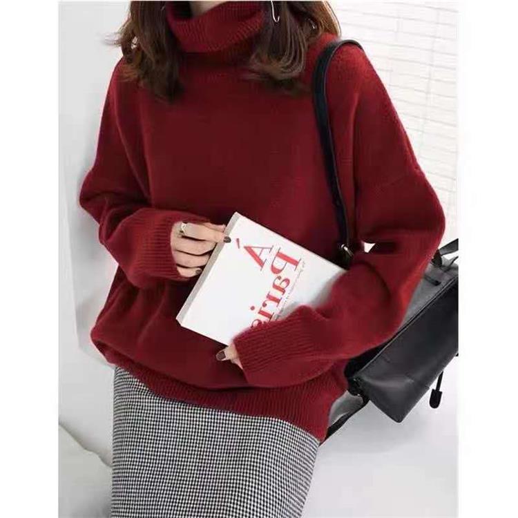 Femme en pull rouge à col roulé tenant un livre, sac noir à l'épaule, jupe à carreaux. Mode automne-hiver, style décontracté élégant.
