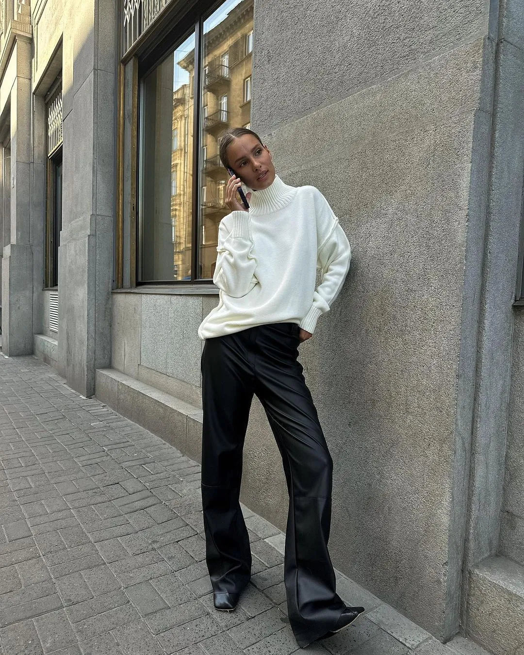 Femme en pull blanc et pantalon noir, posant dans une rue urbaine. Mode automne, look chic et décontracté, architecture moderne en arrière-plan.
