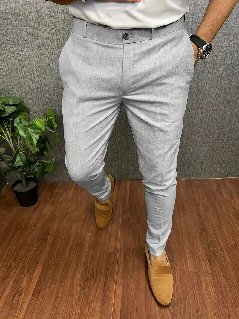 Pantalon gris élégant pour homme, porté avec des mocassins marron clair, sur fond de sol en bois et plante verte. Mode masculine chic.