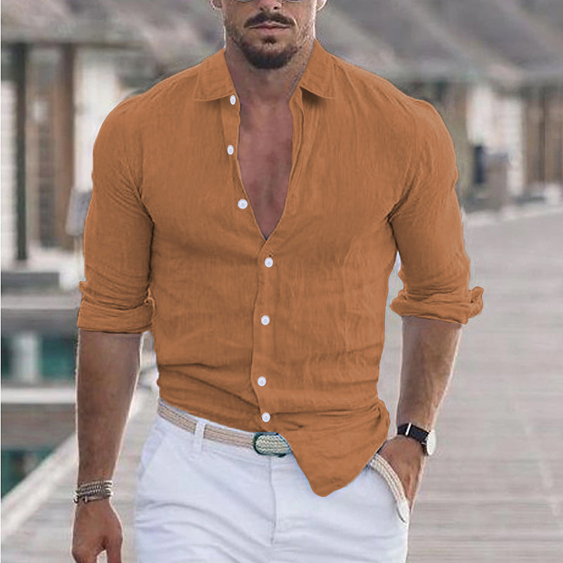 Homme en chemise marron ouverte et pantalon blanc, marchant sur une promenade en bois. Mode masculine, style décontracté, été.