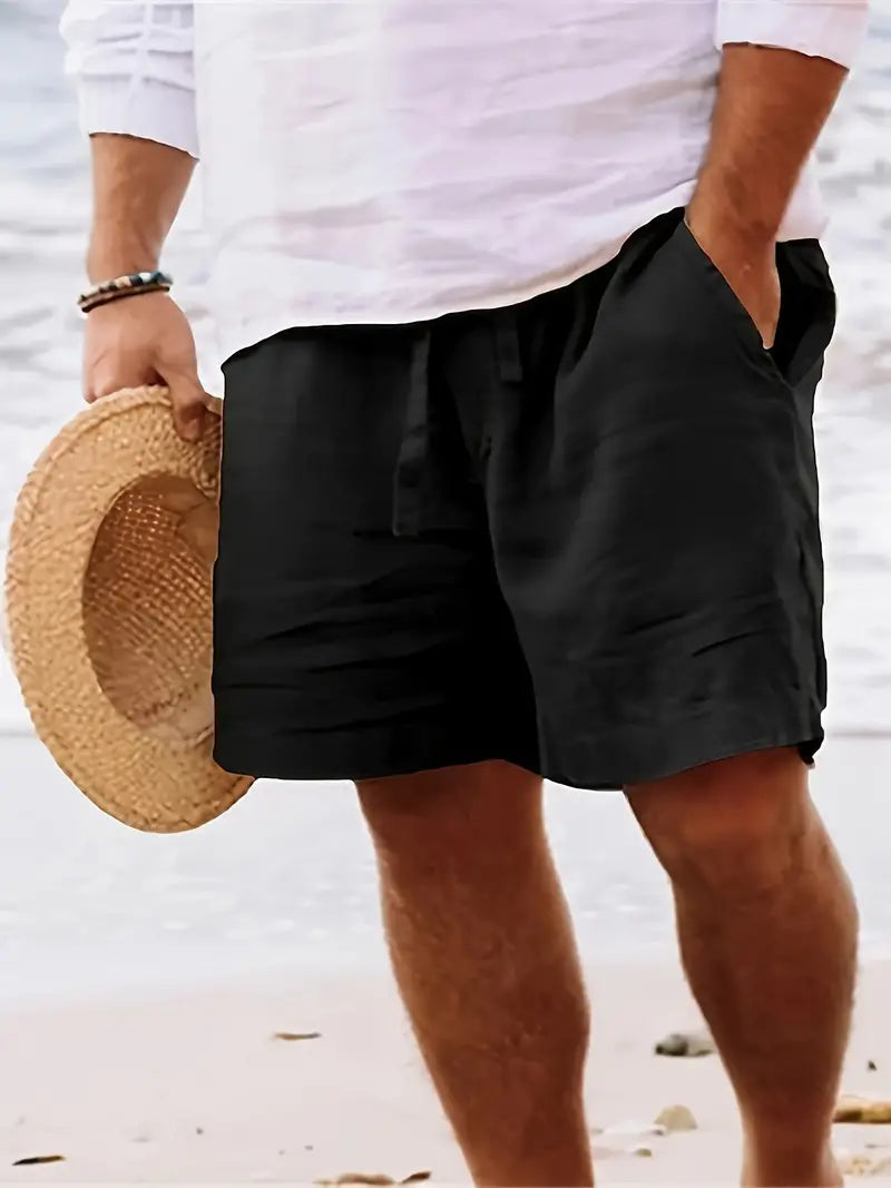 Homme en short noir et chemise blanche tenant un chapeau de paille sur une plage. Mode estivale, tenue décontractée, vacances bord de mer.