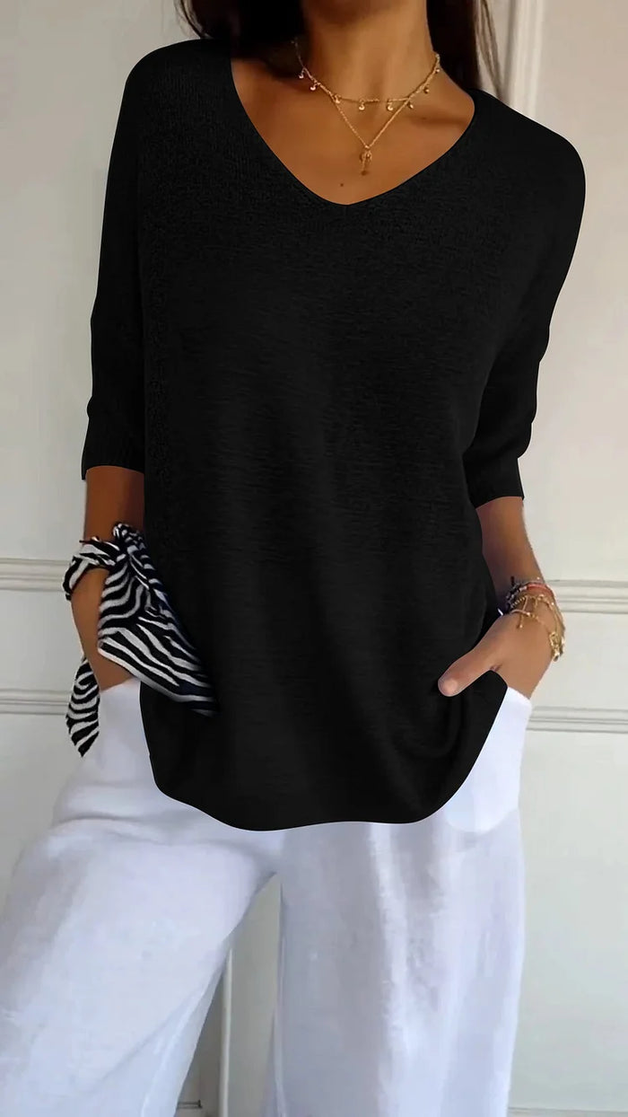 Femme portant un pull noir à col en V, manches retroussées, avec un pantalon blanc. Accessoires: collier doré, bracelet, foulard rayé. Mode élégante.