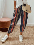 Pantalon rayé multicolore femme, style décontracté, avec sac à main en paille et baskets blanches, sur tapis beige. Mode tendance et confortable.