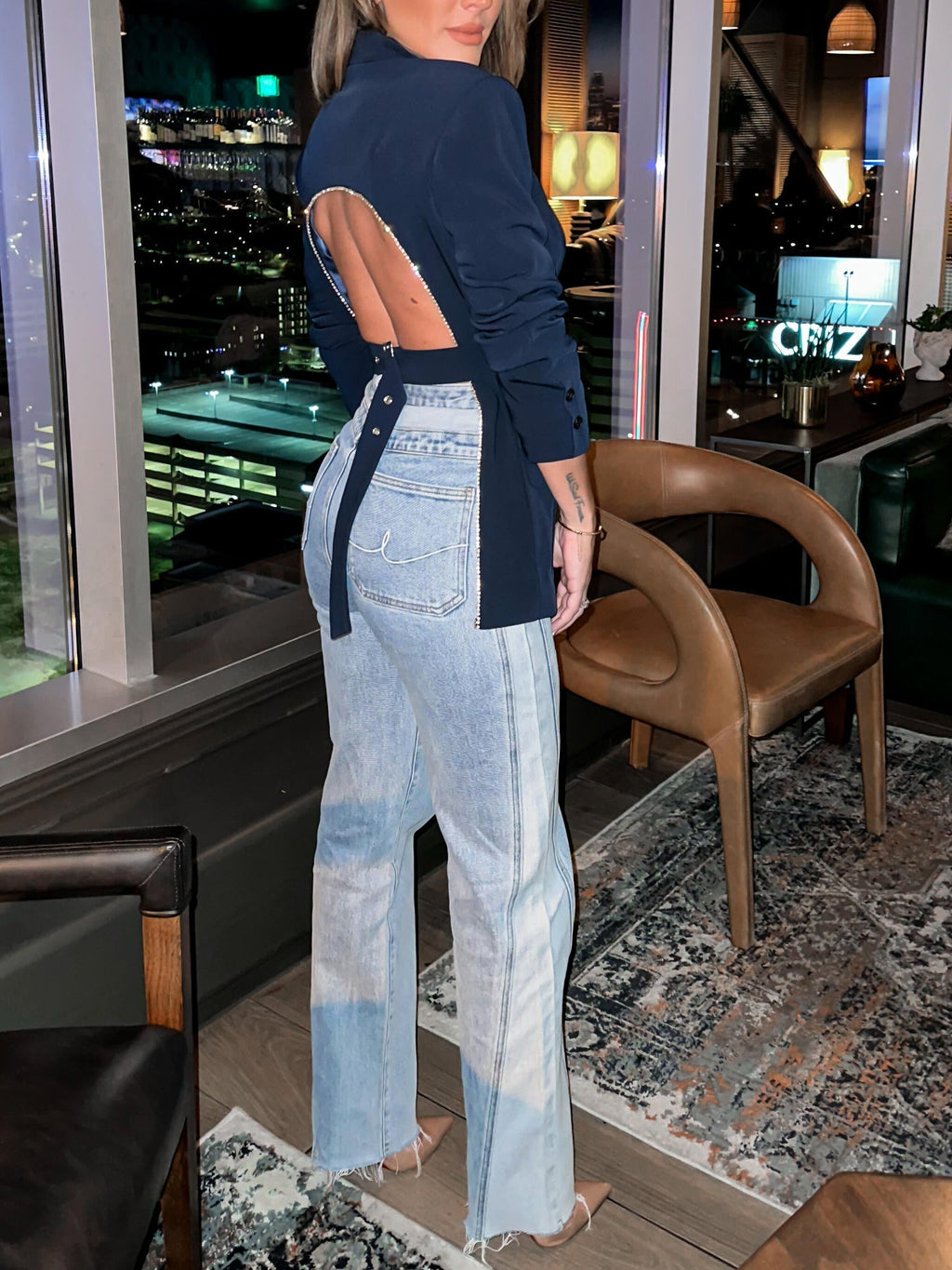 Femme portant un blazer bleu à dos ouvert et un jean délavé, debout dans un salon moderne avec vue nocturne sur la ville. Mode urbaine chic.