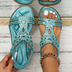 Sandales turquoise ornées de strass, portées sur un pied avec bracelet doré, posées sur un fond en bois blanc. Mode estivale élégante et tendance.